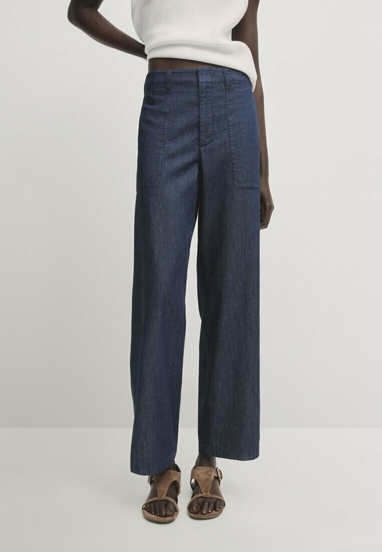 Dunkelblaue Denim-Hosen mit weitem Bein, hoher Taille und Fronttaschen, kombiniert mit braunen Sandalen. Strukturierter Stoff mit glatter Oberfläche.