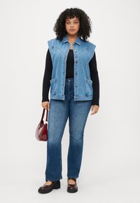 Denim brezrokavnik z gumbom spredaj, prenapolnjenimi armhole in dvema sprednjima žepoma, nošen preko črne dolge majice z dolgimi rokavi in v kombinaciji z razširjenimi kavbojkami.