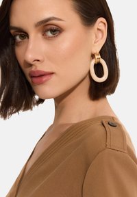 Brązowa bluzka z dekoltem w kształcie litery V, z fakturowanym wykończeniem i guzikiem na ramieniu. Duże kremowe kolczyki w kształcie kół z złotymi akcentami.