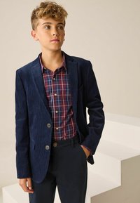 Tumed korduroy bleiser punase ja sinise ruudulise särgi peal, koos tumedate pükstega; silmapaistvad detailid hõlmavad kontrastseid nuppe ja kitsast lõiget.