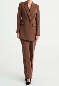 Brun skräddarsydd kavaj med dubbla knappar och sidofickor, tillsammans med matchande smala byxor. Båda har en mjuk textur och rena linjer.