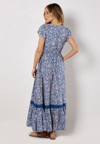 Robe maxi avec un fond bleu, motif floral rose et blanc, taille froncée, manches courtes et ourlet à plusieurs niveaux. Présente un accent bleu uni en bas.