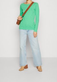 Maglietta verde a maniche lunghe in materiale leggero, abbinata a jeans a gamba larga di colore blu chiaro e stivaletti con tacco testurizzati color beige. Inclusa borsa a tracolla marrone.
