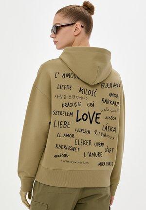 Persoon draagt een beige hoodie met "love" geschreven in meerdere talen op de rug, gecombineerd met bijpassende broek en zonnebril, haar in een knot.