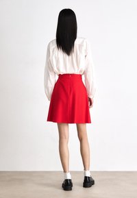 Jupe évasée rouge à texture lisse ; fermeture éclair au dos. Associée à un chemisier blanc à manches longues et des chaussures noires épaisses.