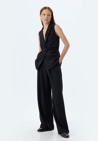 Svart ärmlös jumpsuit med djup V-halsringning, bälte i midjan, vid byxben och mjukt tyg. Slät struktur med skräddarsydda detaljer.