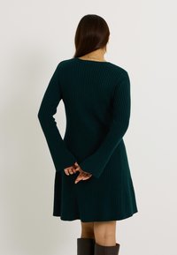 Robe côtelée vert foncé avec de longues manches évasées et un ourlet évasé, présentant un large décolleté et une texture lisse, vue de dos.