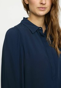 Blouse bleu marine avec un col, une patte de boutonnage et des détails de manches froncées, fabriquée dans un tissu lisse et léger.