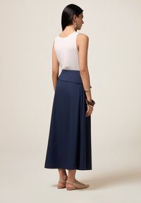 Navy blauwe maxi rok met een aansluitende taille, gemaakt van een gladde stof. Gecombineerd met een wit tanktop en beige ballerina's, zichtbaar vanuit de achterkant.