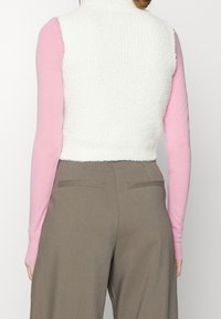 Top senza maniche bianco testurizzato stratificato su una camicia a maniche lunghe rosa, abbinato a pantaloni beige a vita alta con tasche posteriori.