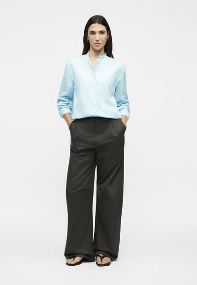 Γυναίκα στέκεται με τα χέρια στις τσέπες φορώντας ένα ανοιχτομπλε πουκάμισο με κουμπιά, φαρδιά μαύρη παντελόνα και μαύρες σαγιονάρες σε λευκό φόντο.