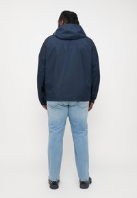 Marine vanntett jakke med justerbar hette, elastiske mansjetter og en avslappet passform, kombinert med lyseblå jeans og svart fottøy.