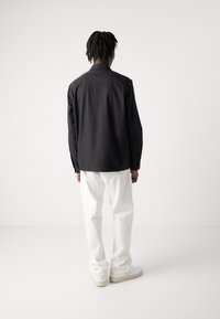 Camisa negra de manga larga con cuello, combinada con pantalones blancos, que muestra un ajuste relajado y una textura suave, con sencillos detalles de botones.