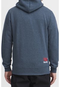 Uomo che indossa una felpa con cappuccio di colore blu scuro con il logo rosso e bianco "SLD Since 1993" nella parte bassa della schiena, in piedi con le braccia rilassate.