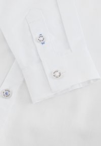 Poignet de chemise blanche avec deux boutons visibles gravés "Baker by Ted Baker" et fil bleu maintenant les boutons.