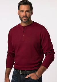 Burgunderfarbendes Longsleeve-Henley-Shirt mit drei Knöpfen, gerippter Textur und Rundhalsausschnitt. Kombiniert mit dunklen Jeans und einem schwarzen Gürtel.