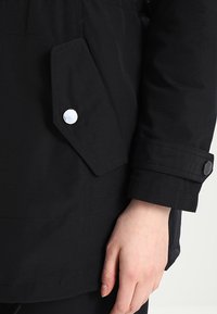 Chaqueta negra con un tejido texturizado, bolsillo en ángulo con un botón a presión plateado y un detalle de puño en contraste.