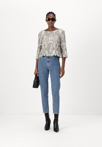 Beiger Spitzenoberteil mit floralen Mustern, ausgestellten Ärmeln, blauen hochgeschnittenen Jeans und schwarzen, absatzlosen Stiefeletten; hält eine kleine schwarze Tasche.