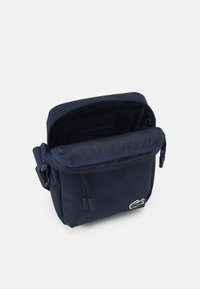 Bolso bandolera azul marino hecho de un tejido resistente, que cuenta con un compartimento principal con cierre, un bolsillo frontal y un acento de logo bordado.
