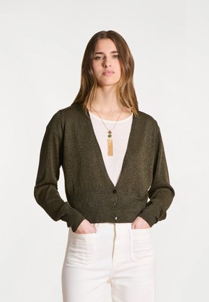 Jeune femme portant un cardigan court olive foncé sur une chemise blanche, un pantalon blanc et un long collier pendentif, les mains dans les poches.