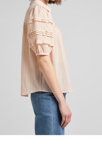 Blusa color pescozza con finitura testurizzata, dotata di accenti arricciati sulle maniche corte e collo a punta. Indossata con jeans in denim blu.