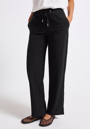 Zwarte, lichte, elastische broek met een trekkoord in de taille, voorzien van zijzakken en een losse pasvorm. Gecombineerd met open teen zwarte mesh schoenen.