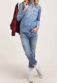 Person klädd i en denimskjorta och broderade jeans med uppvikta manschetter, vita sneakers och håller en röd och svart rutig jacka över ena axeln.