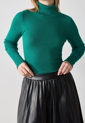 Kobieta ubrana w dopasowany zielony prążkowany golf i czarną plisowaną skórzaną spódnicę, poprawiająca pas spódnicy.