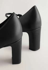 Pantofi negri cu toc înalt, realizați din piele netedă; au vârf pătrat și toc gros, cu o strălucire subtilă și linii curate.