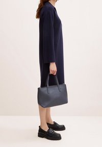 Borsa a tracolla in pelle nera con una texture, dotata di due manici robusti, che si abbina a un abito blu scuro lungo fino al ginocchio. Modello che indossa mocassini neri.