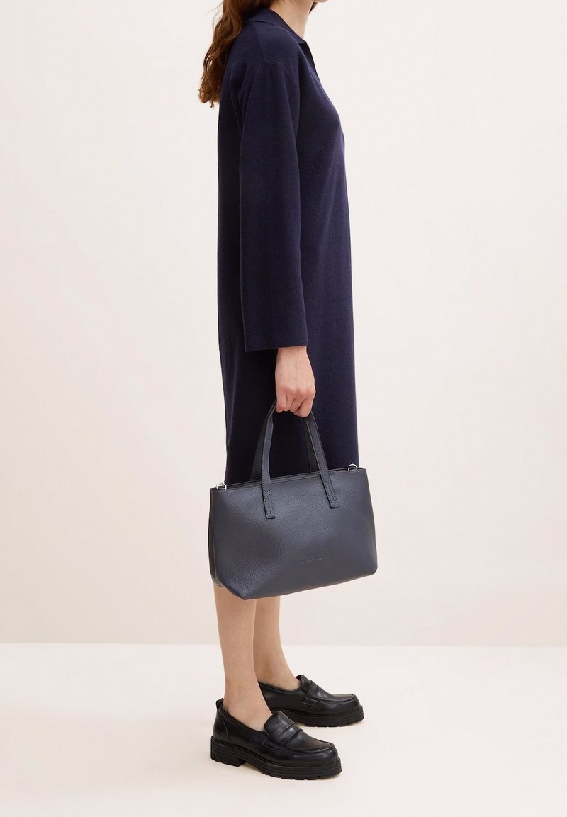 Borsa a tracolla in pelle nera con una texture, dotata di due manici robusti, che si abbina a un abito blu scuro lungo fino al ginocchio. Modello che indossa mocassini neri.