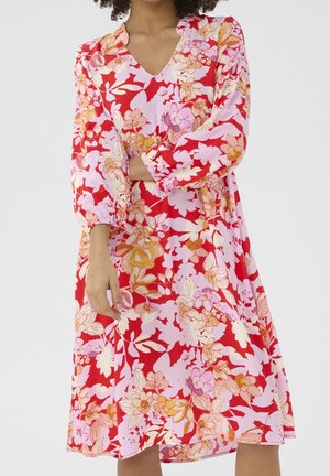 Vrouw draagt een rode jurk met roze en oranje bloemenpatronen en lange opgeblazen mouwen met elastische manchetten.