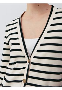 Pasiasty kardigan w kolorze ecru i czarnym, z dekoltem w serek, złotymi guzikami i miękką teksturą dzianiny. Widoczne nakładanie na białą bluzkę.