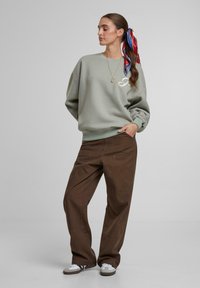 Lys grøn genser med rund hals og oversized ermer, kombinert med løse brune bukser. Hvite detaljer og et mønstret hodebånd fullfører stilen.