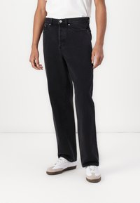 Black denim jeans with a straight leg cut, featuring two front pockets, a button fly, and silver-tone hardware. Worn with white sneakers.