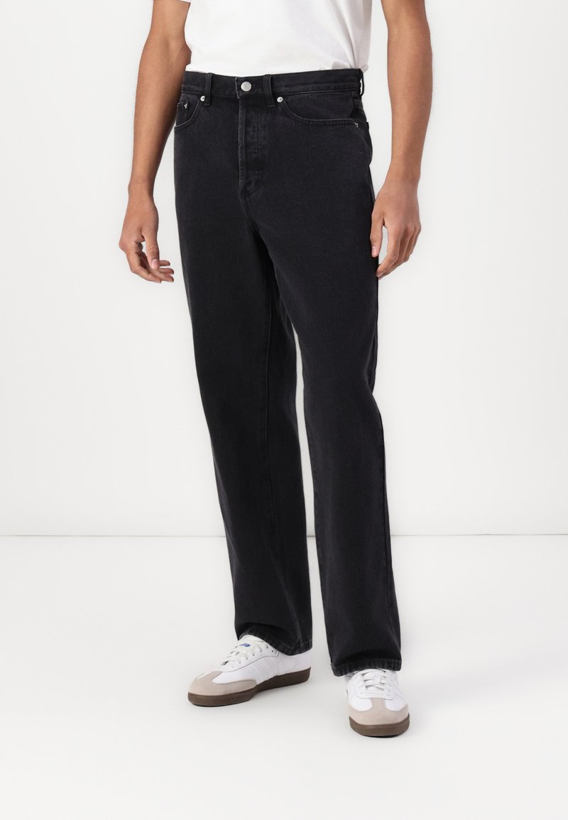 Black denim jeans with a straight leg cut, featuring two front pockets, a button fly, and silver-tone hardware. Worn with white sneakers.