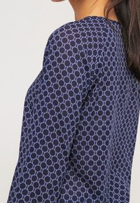 Mörkblå blus med ett cirkulärt mönster i ljusare blått, med rund halsringning och långa ärmar. Mjuk, lätt tyg.