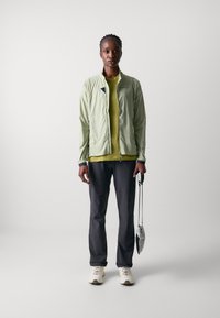 Veste zippée verte clair sur un haut jaune, associée à un pantalon foncé et des baskets blanches. Tenant un petit parapluie gris.