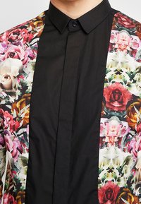 Chemise à manches courtes avec devant noir et motif floral coloré sur les épaules, comprenant des roses et d'autres fleurs, tissu texturé.