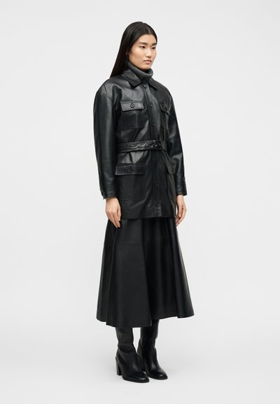 Γυναίκα που φοράει μαύρο δερμάτινο μπουφάν με ζώνη, ταιριαστό δερμάτινο midi φούστα, μαύρες μπότες με τακούνι και γκρι πουλόβερ με ψηλό γιακά.