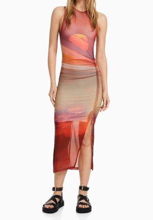 Kobieta w sukience midi bez rękawów z nadrukiem zachodu słońca, z marszczeniem po boku, rozcięciem z boku oraz czarnymi platformami zdobionymi ćwiekami.