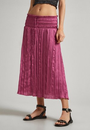 Kvinde iført en højtaljet, plisseret nederdel i ankellængde i magenta med sorte flade sandaler og en sort cropped top.