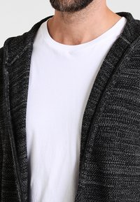 Cardigan nero texture con cappuccio, caratterizzato da una vestibilità ampia, indossato su una maglietta bianca semplice. Dettagli di cucitura visibili lungo i bordi.