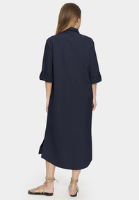 Rochie albastru marin, până la genunchi, cu guler, mâneci suflecate și slituri laterale. Textură netedă, realizată dintr-un material ușor. Accente de sandale vizibile.