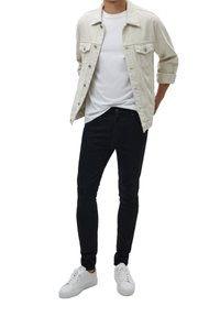 Chaqueta de mezclilla beige claro con botones delanteros, camiseta blanca de cuello redondo, jeans ajustados negros y zapatillas blancas.