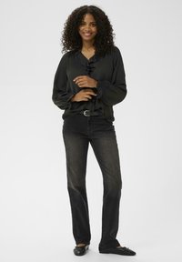 Blusa nera a volant con maniche lunghe, abbinata a jeans skinny scuri e ballerine nere. Il modello è in piedi contro uno sfondo neutro.