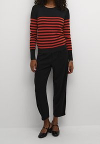 Suéter negro con rayas horizontales rojas, combinado con pantalones negros y zapatos de charol negros. El conjunto presenta mangas largas y un cuello redondo.