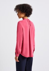Camicia di seta rosa con maniche lunghe, colletto a volant texturizzato e polsini arricciati. Sul retro presenta una piega sottile per un movimento aggiuntivo.