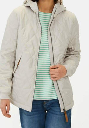 Kvinde iført lysebeige quiltet jakke over grøn- og hvidstribet skjorte og blå jeans, der holder i jakkezippen.