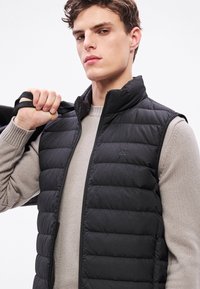 Μαύρο μπουφάν τύπου puffer με φερμουάρ, ψηλό γιακά και μπροστινές τσέπες. Φορεμένο πάνω από ανοιχτό γκρι πλεκτό πουλόβερ, με ραμμένη υφή και λεπτομέρεια λογότυπου.